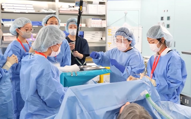 手術室で手術台の脇に立ち、器具の使い方を学ぶ（洛和会音羽病院ご提供）